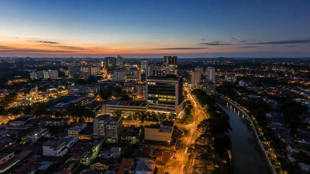 Dusk aerial across a Malaysian urban corridor of high-rise towers, highways, shophouses and industrial sheds